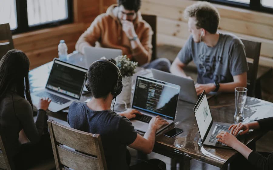 Five people working on laptops around a wooden table, one wearing headphones with code on screen